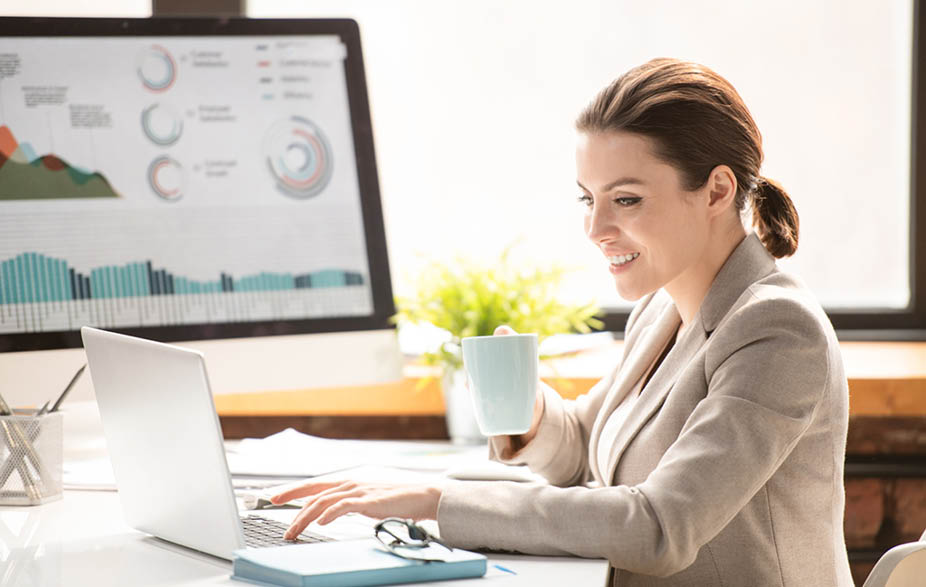 Happy young female broker with mug having tea while sitting in front of laptop and communicating online at break