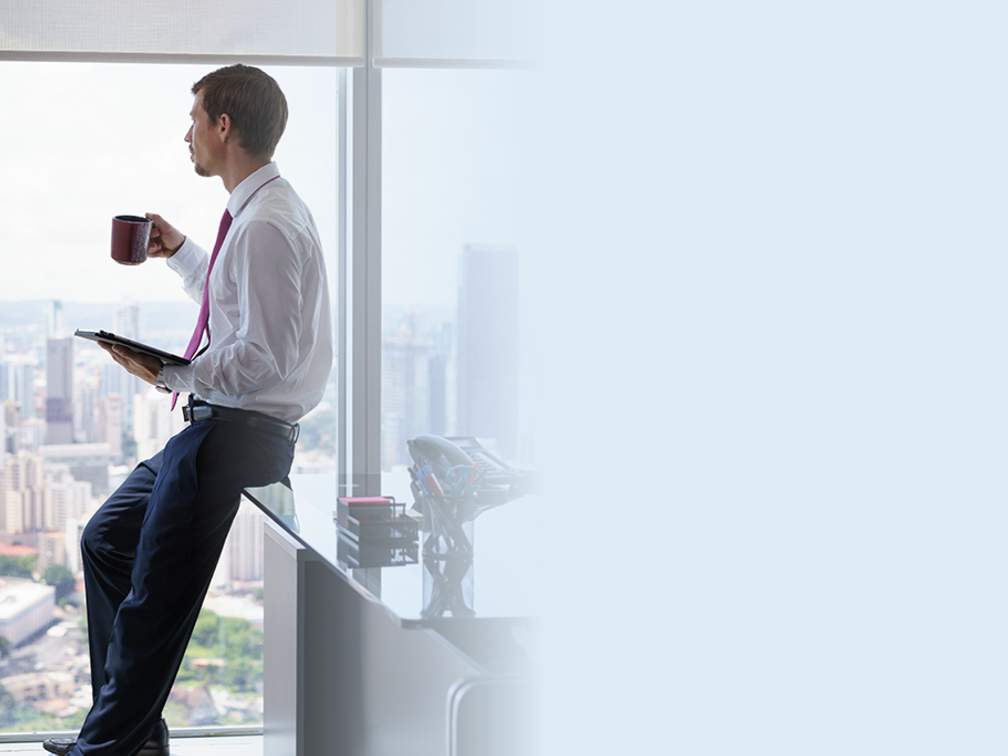 Adult businessman sitting on desk in modern office and reading news on tablet pc with a cup of coffee  The man looks out of the window and contemplates the city and skyscrapers 