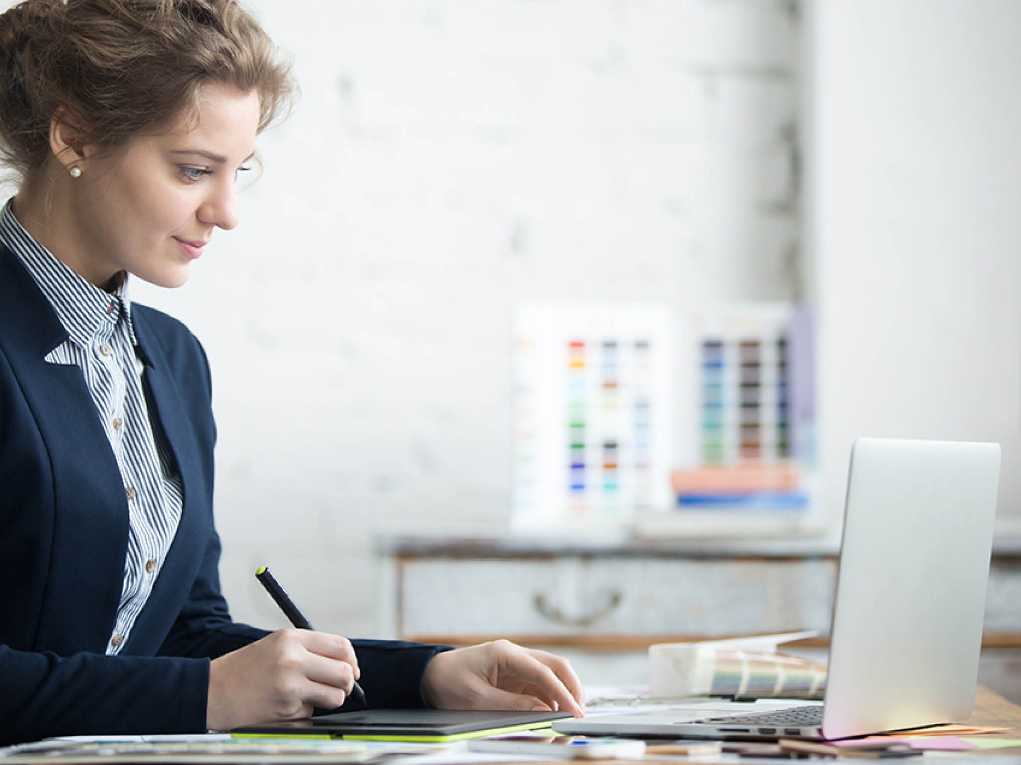 Portrait of beautiful happy young graphic designer woman working at home office desk  Attractive model wearing suit using digital tablet, sitting in front of laptop  Interior shot